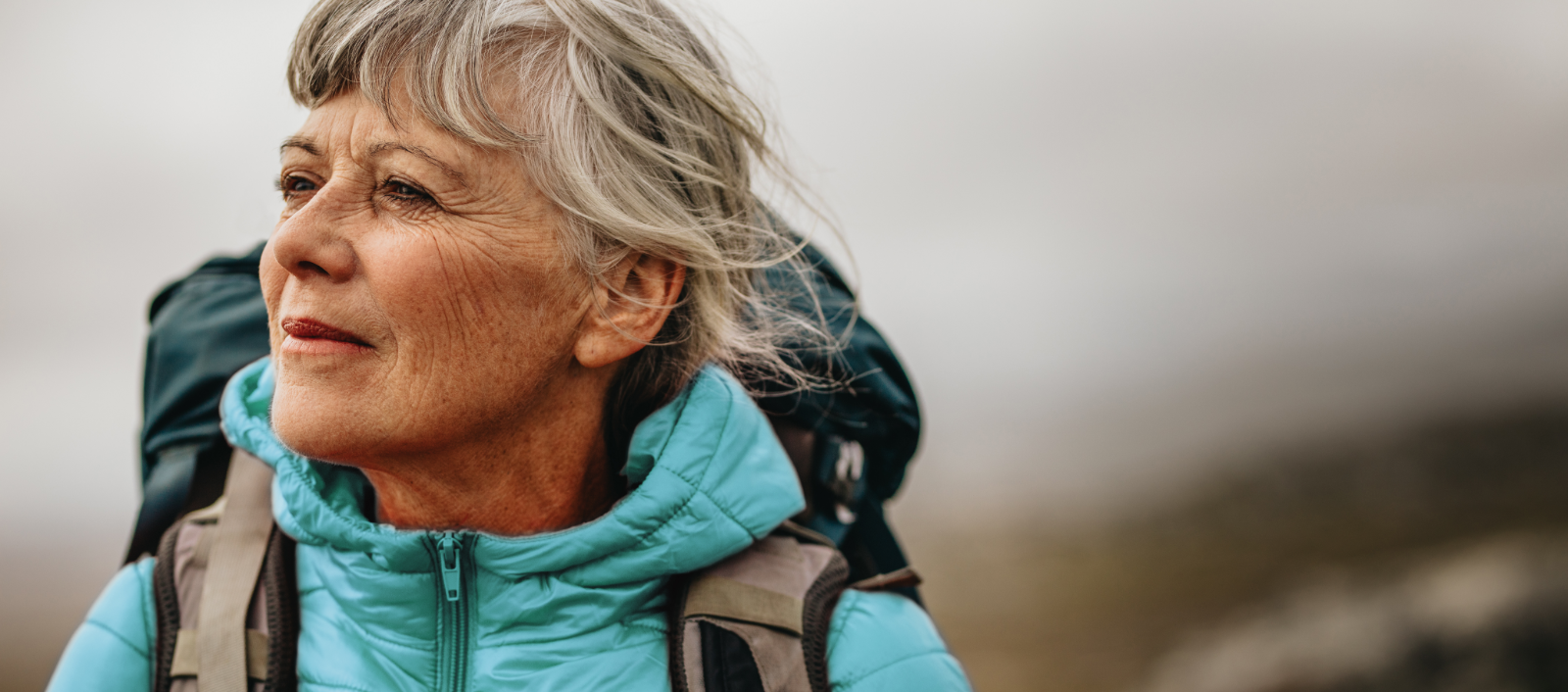 an elder woman with gray and white hair is wearing a blue coat and teal backpack with gray straps while gazing off to the left.