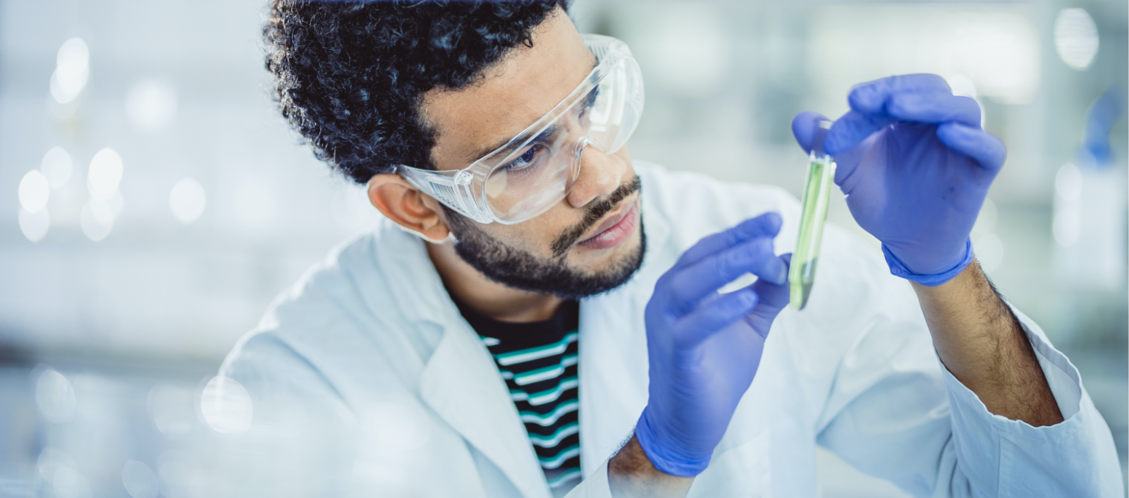 Man with curly black hair and beard wears a striped shirt under his white lab coat, safety googles, and blue latex gloves while examining a test tube of transparent green liquid.